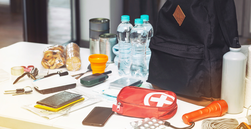 Assortment of hurricane preparedness items on a desk: flashlight, canned goods, water, medication, documents, phone, backpack, and more.